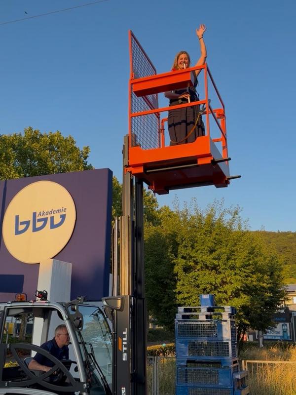 Mitarbeiterin auf Hebebühne winkt vor dem bbv Akademie Gebäude